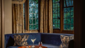 Un canapé bleu agrémenté de coussins à motifs, une table ronde en bois sur laquelle reposent un verre à martini et un verre à vin, et une vue sur la verdure à travers de grandes fenêtres ornées de rideaux.