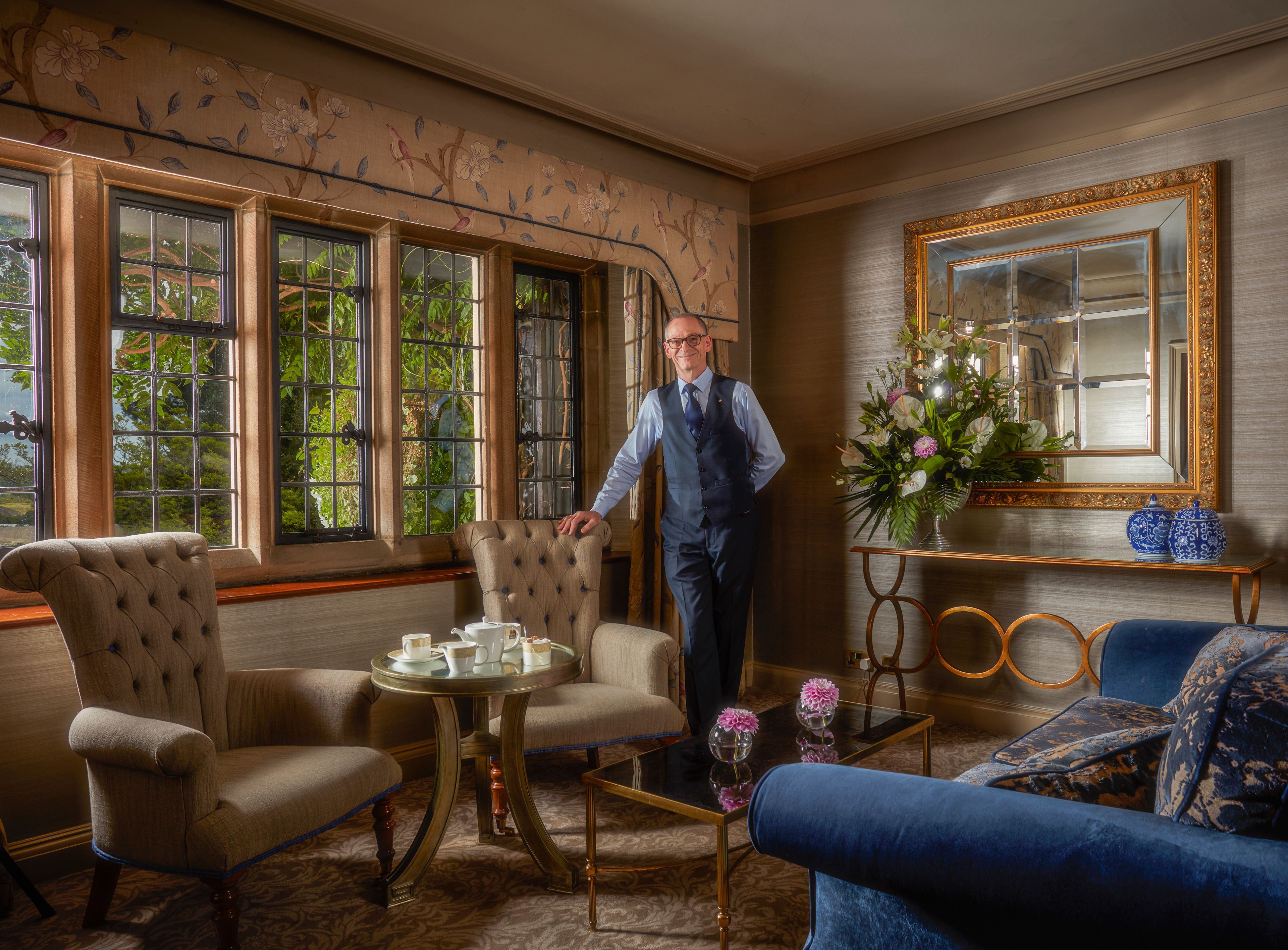 A room with a large window, a mirror, a blue sofa, and a table with a vase of flowers.