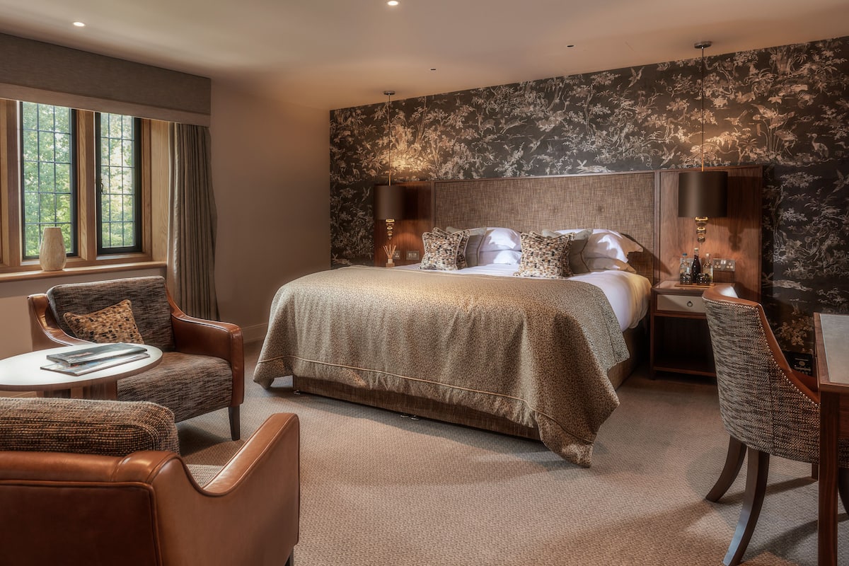 Habitación de hotel moderna con una cama grande, dos sillas, una mesita y una mesita de noche con lámpara.