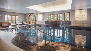 Un espace piscine intérieur moderne, doté d’une paroi en verre, de chaises longues et d’une verrière.