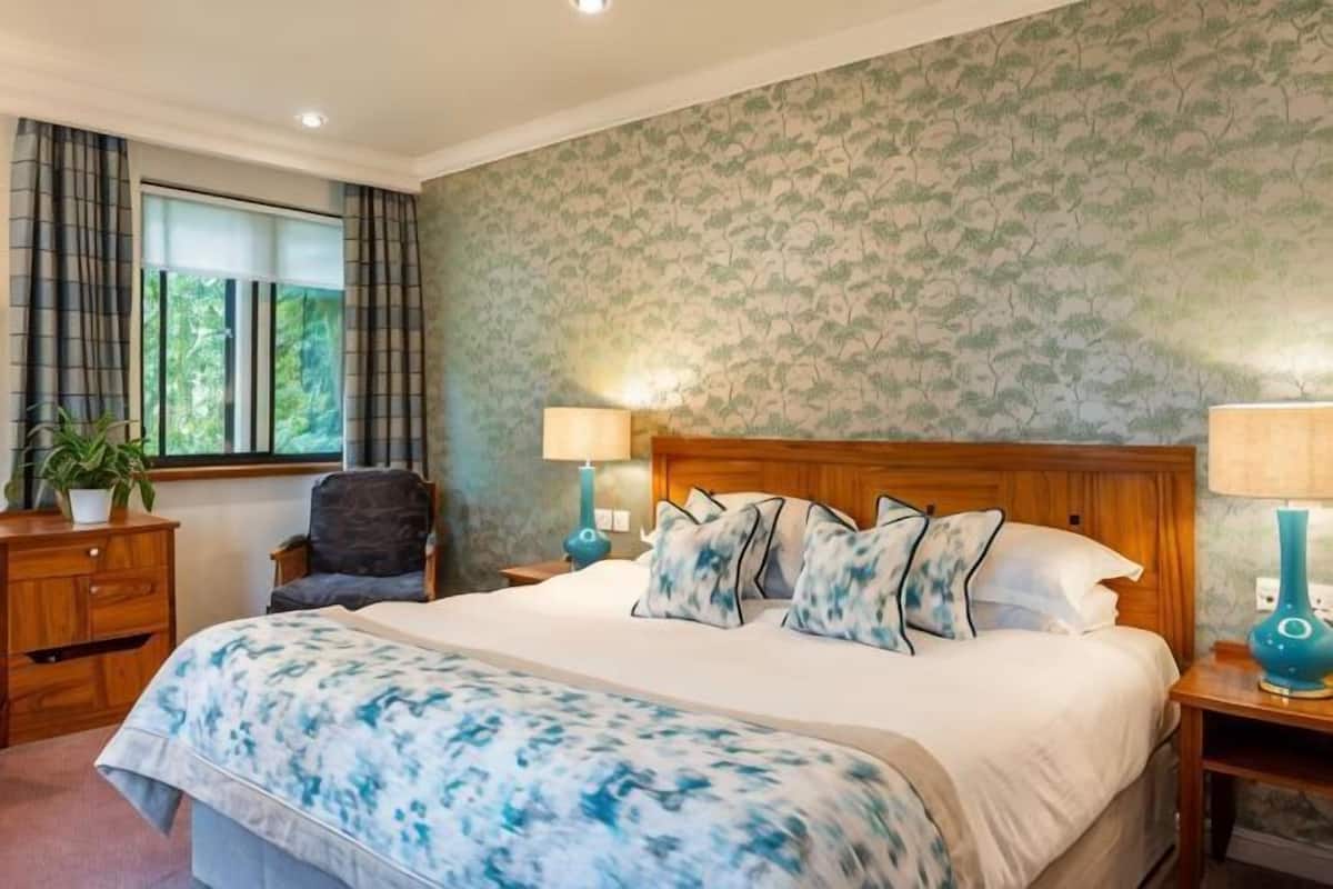 Un dormitorio con una cama grande, dos mesitas de noche con lámparas, una silla, un ventanal con cortinas y una planta.