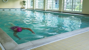 An indoor swimming pool with clear water, a swimmer in a pink swimsuit, and large windows allowing natural light.