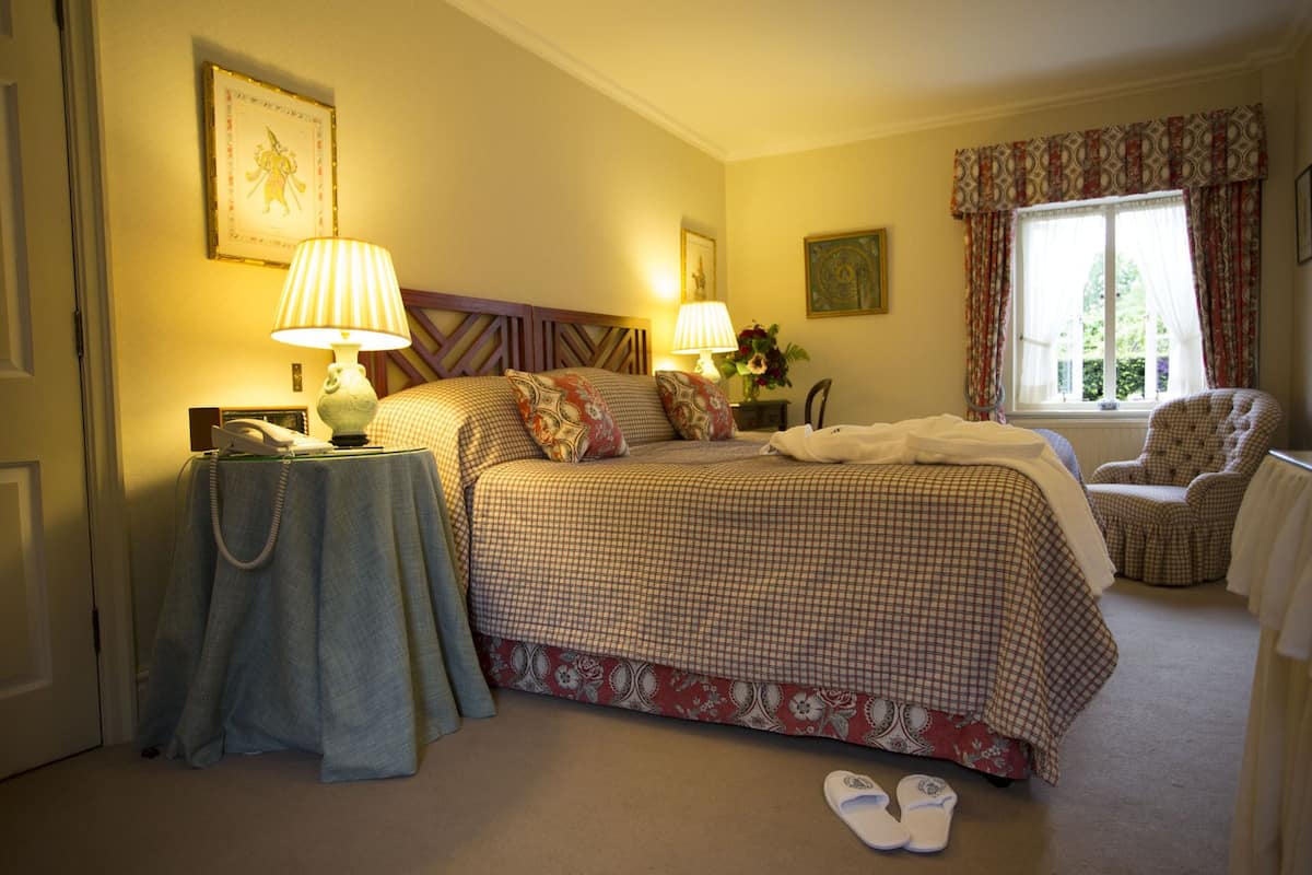 A neatly arranged bedroom with a checkered bedspread, two bedside lamps, a chair, and a window with curtains.