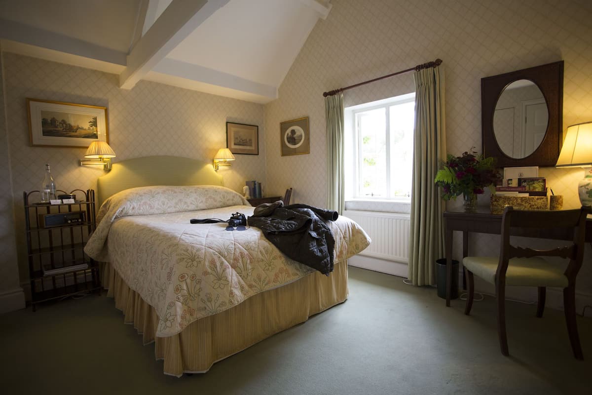 A bedroom with a bed, bedside lamps, a desk with a chair, a window with curtains, and a mirror.