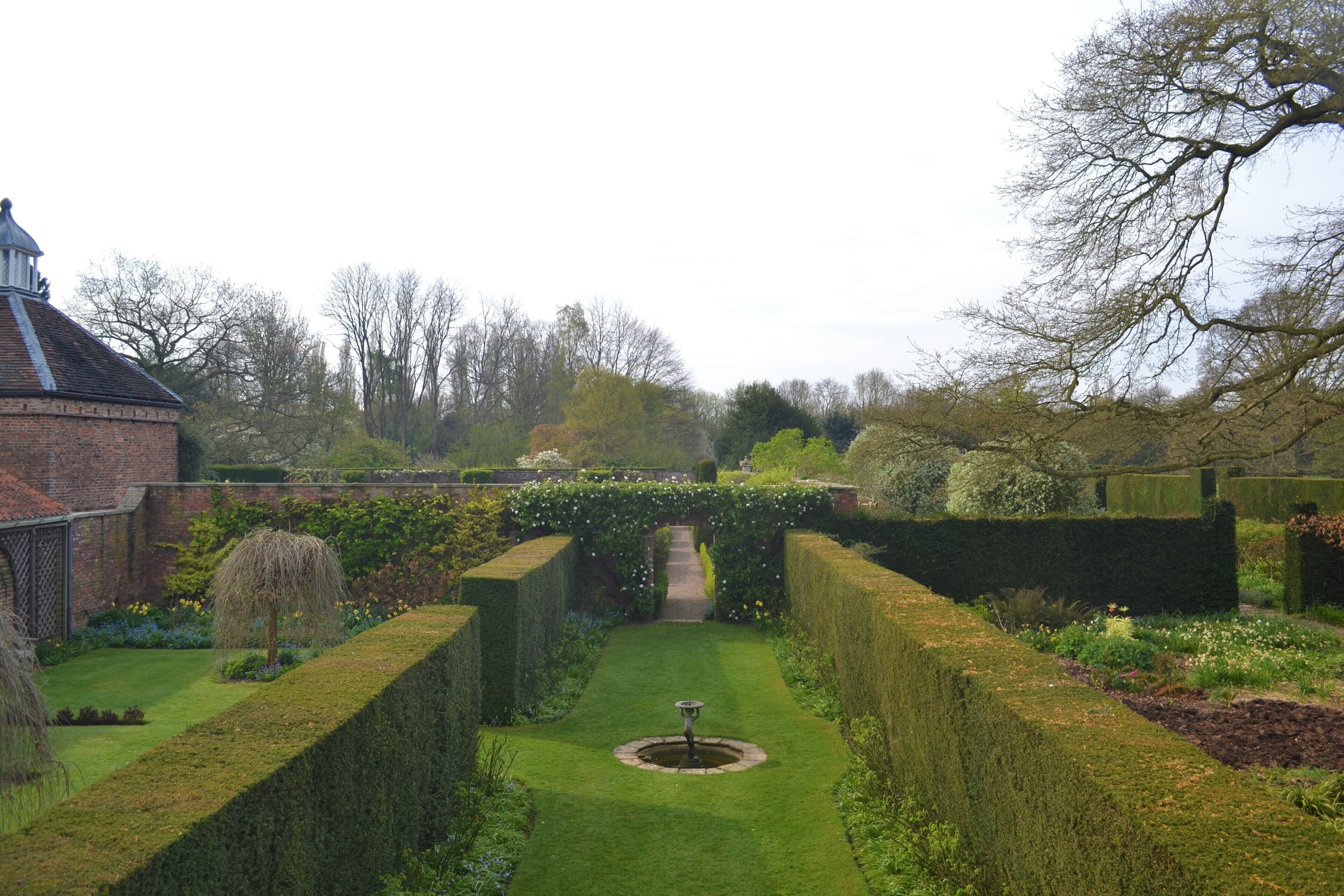 A formal garden with a central fountain, neatly trimmed hedges, and a brick building with a dome.