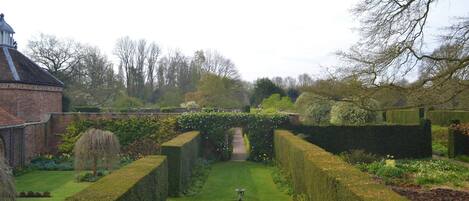 A formal garden with a central fountain, neatly trimmed hedges, and a brick building with a dome.