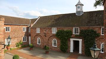 A courtyard with a brick building featuring arched windows and a central door, surrounded by potted plants and a gravel path.