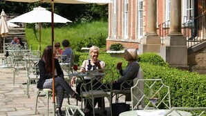 An outdoor café with tables and chairs, surrounded by greenery and a building with columns.
