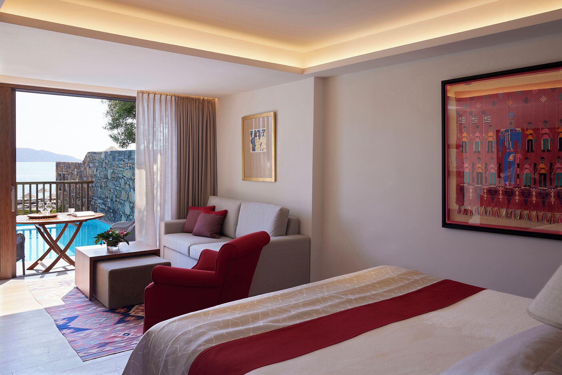 Una habitación de hotel con una cama, un sofá, un sillón rojo, una mesita con una bandeja y vistas a una piscina y montañas.