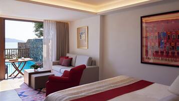 Una habitación de hotel con una cama, un sofá, un sillón rojo, una mesita con una bandeja y vistas a una piscina y montañas.