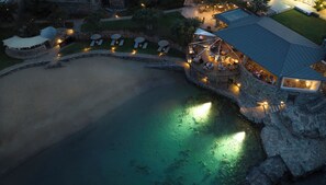 An aerial view of a coastal resort with a large sandy beach, a pool, and a dining area with outdoor seating. - Elounda Mare Relais & Chateaux Hotel (Agios Nikolaos)