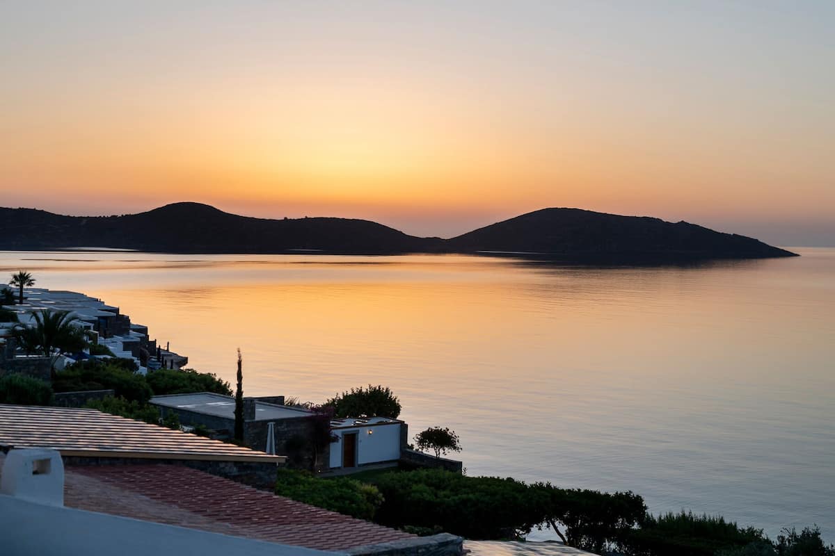 Blick auf die Küste mit Sonnenuntergang über ruhigem Wasser, einer kleinen Insel und einem Gebäude mit roten Ziegeldach.