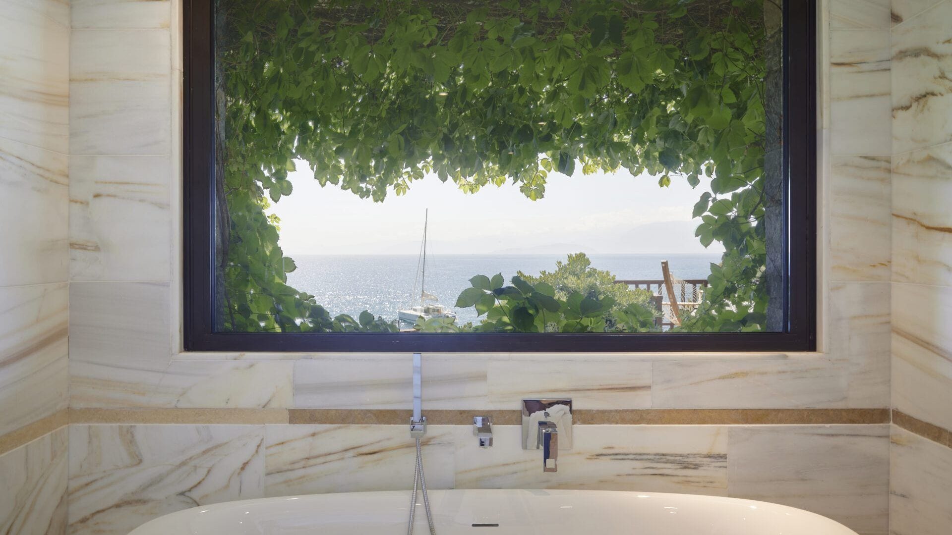 Un baño con piso de mármol, una bañera, un ventanal con vista al mar y una terraza de madera afuera.