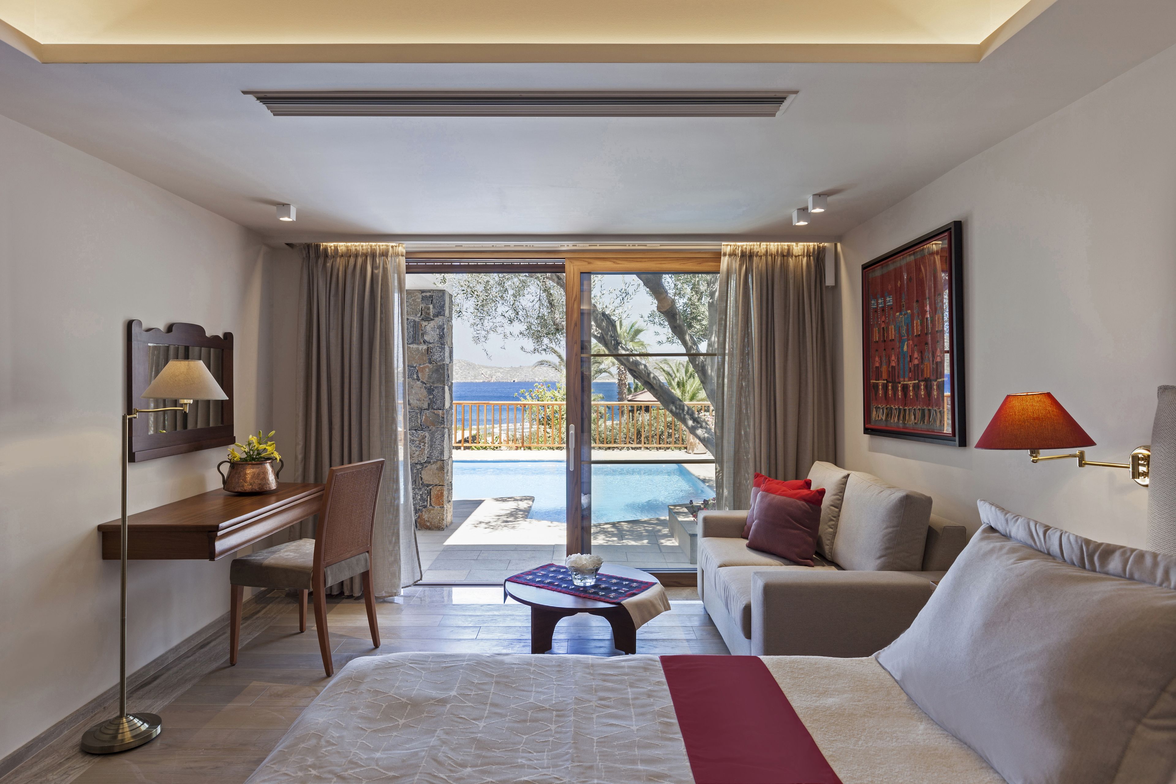 A hotel room with a bed, a desk, a chair, a sofa, a table with a vase, a painting, and a view of a pool and outdoor area.