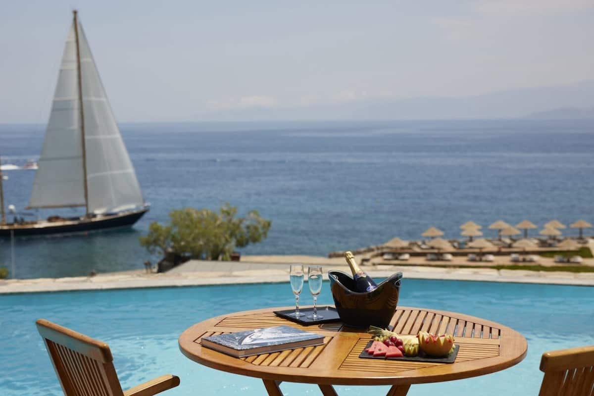 Ein Tisch am Pool mit Buch, Obst, Champagner und Gläsern, mit Blick auf einen Strand mit Sonnenschirmen und einem Segelboot.