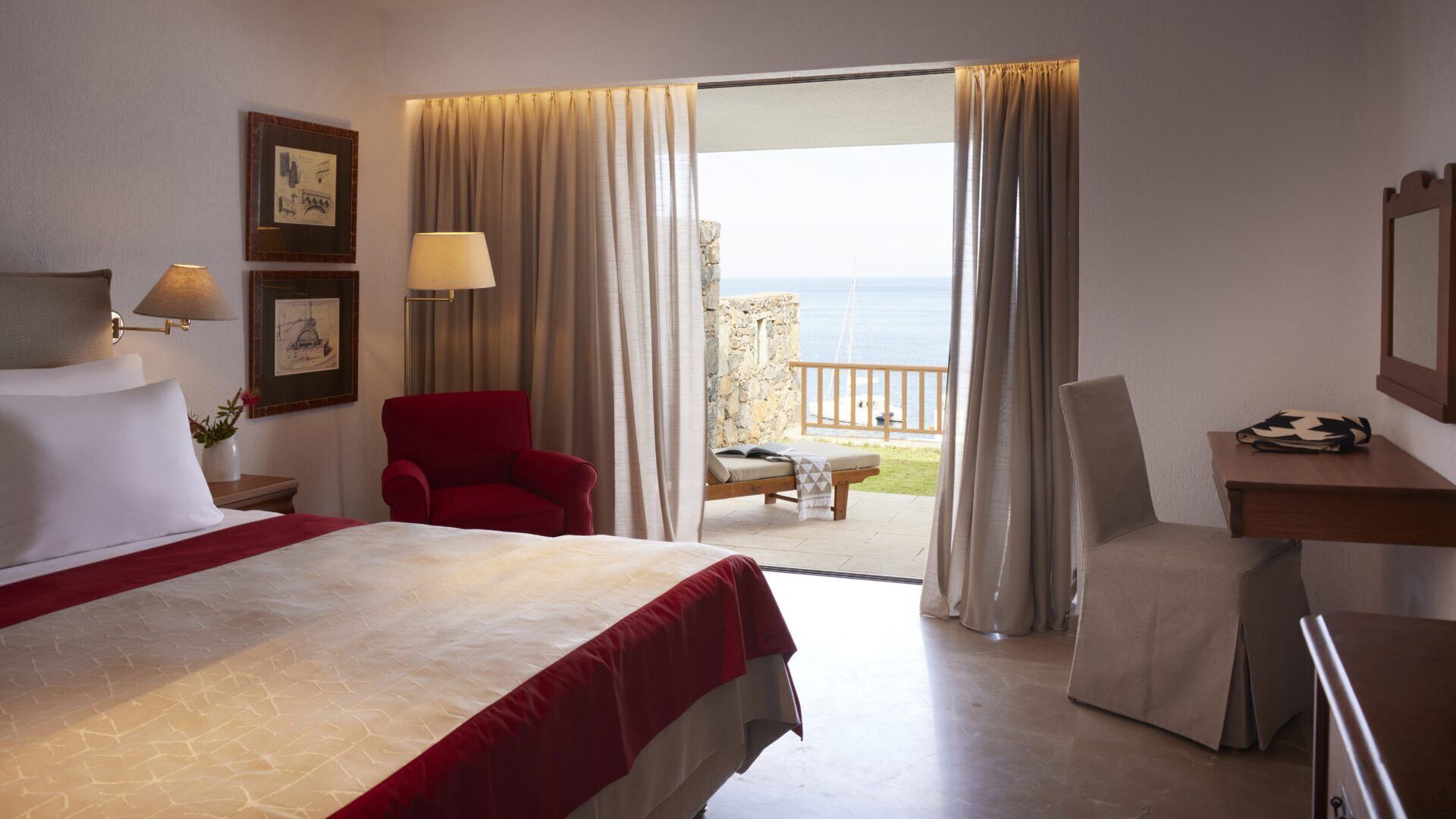 A hotel room with a bed, two chairs, a desk, a lamp, and a view of the sea through a balcony.