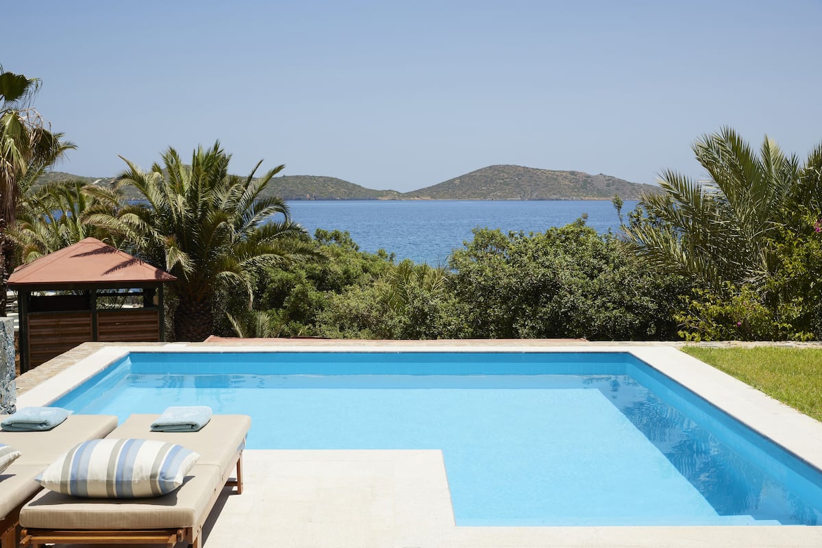 Ein rechteckiger Swimmingpool mit Liegestühlen und Handtüchern, umgeben von Grünpflanzen und Blick auf das Meer und die Hügel.