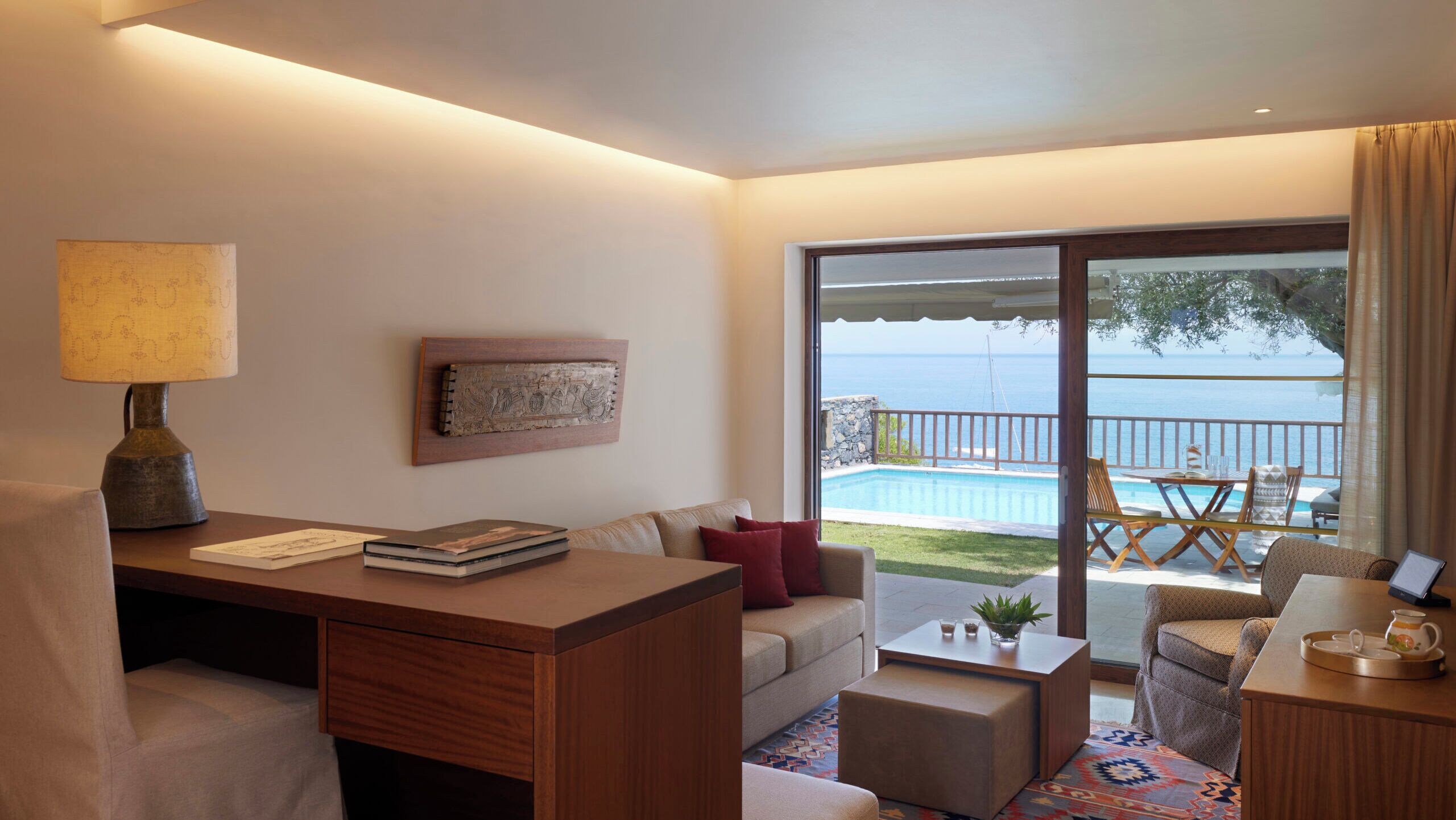 Una habitación de hotel moderna con escritorio, sofá y sillón, un ventanal con vistas al mar y una pequeña zona de piscina.