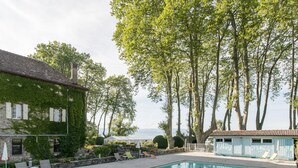 Une piscine entourée de chaises longues, un bâtiment en pierre recouvert de lierre et, au fond, de grands arbres.