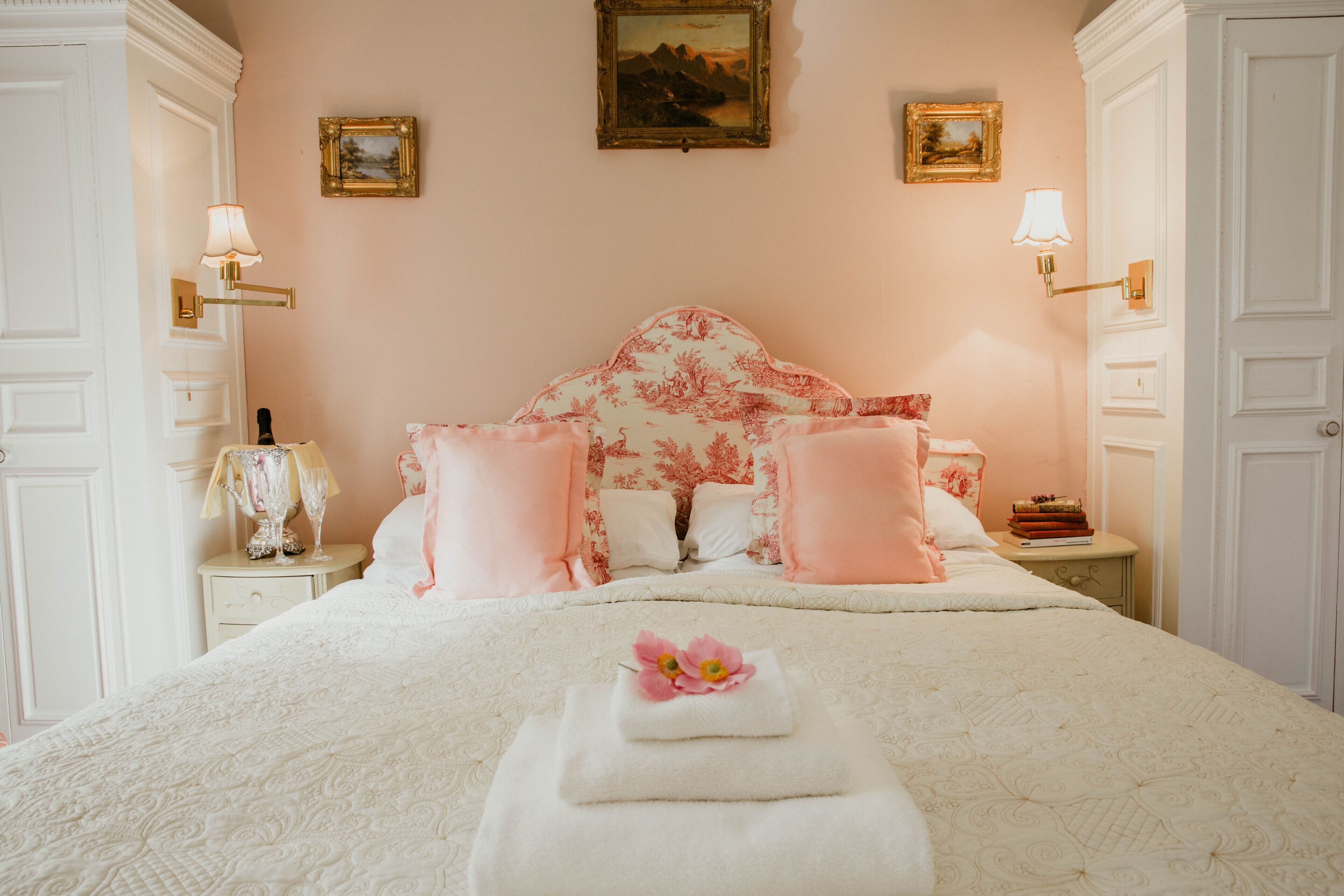 Ein Schlafzimmer mit einem blumigen Kopfteil, einem ordentlich bezogenen Bett mit rosa Kissen, einem Nachttisch mit Lampe und einem gerahmten Bild an der Wand.