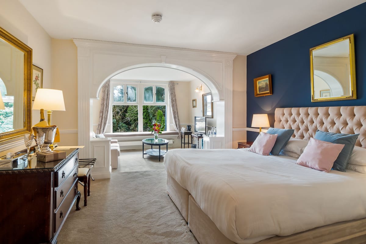Ein Hotelzimmer mit einem großen Bett, einer Sitzecke mit Sofa und Sesseln, einem Esstisch mit Stühlen und Blick auf Grünflächen durch das Fenster.