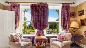 Une pièce avec deux fauteuils, une petite table et une lampe. Les fenêtres sont dotées de rideaux, et on peut admirer un jardin depuis l’intérieur.