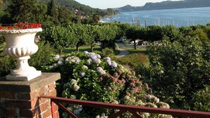 Un giardino con un vaso in pietra, vegetazione rigogliosa e vista su un lago con barche.