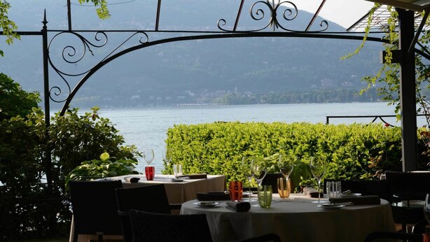 Un ristorante con posti a sedere all'aperto affacciato su un lago, con tavoli apparecchiati con bicchieri da vino e vista sull'acqua.