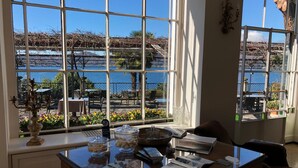 Camera d'albergo con un'ampia finestra che si affaccia su una spiaggia e il mare. Si trova una scrivania con un vaso, una ciotola di biscotti e alcune riviste.