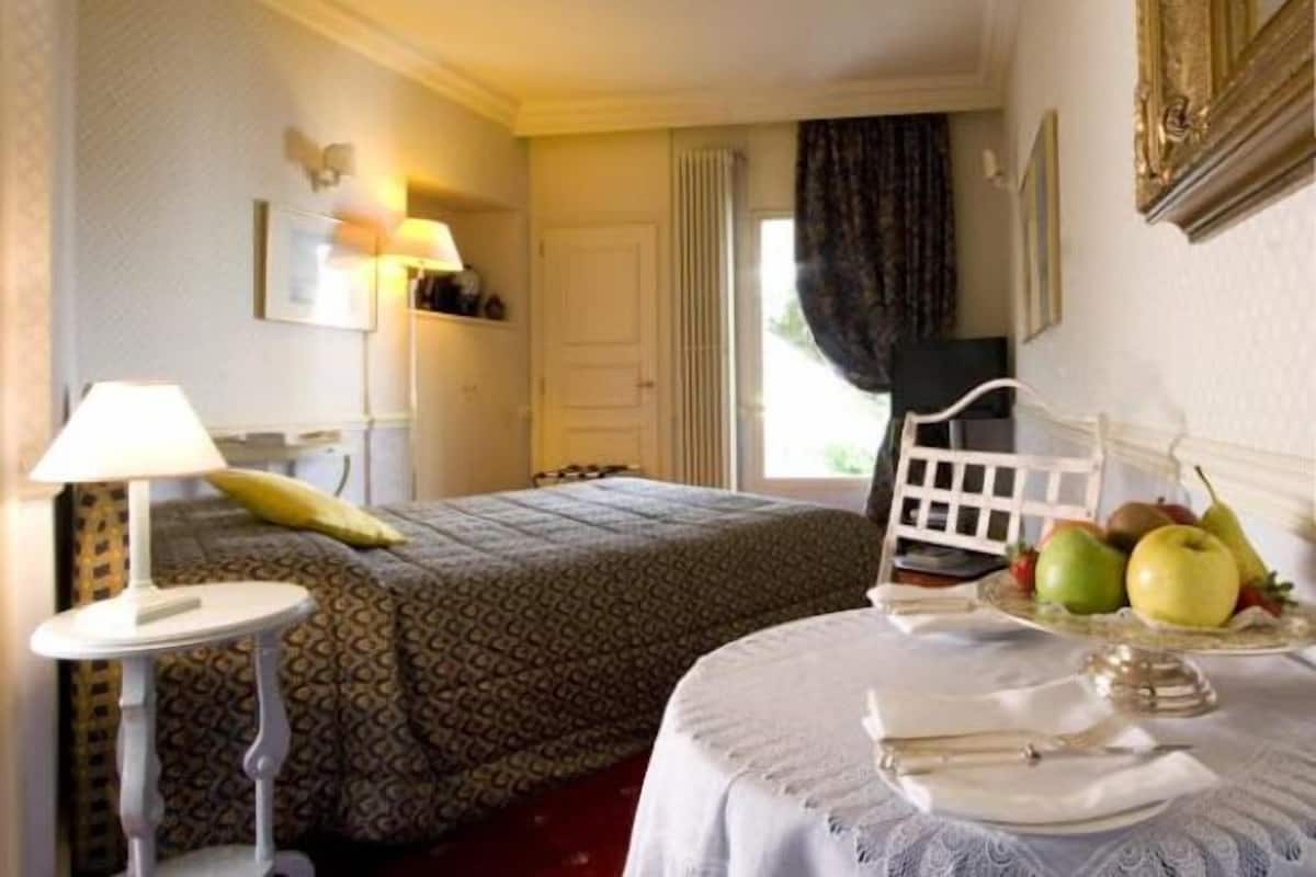 A hotel room with a bed, a bedside table with a lamp, a chair, and a table with fruits and a tray of cutlery.