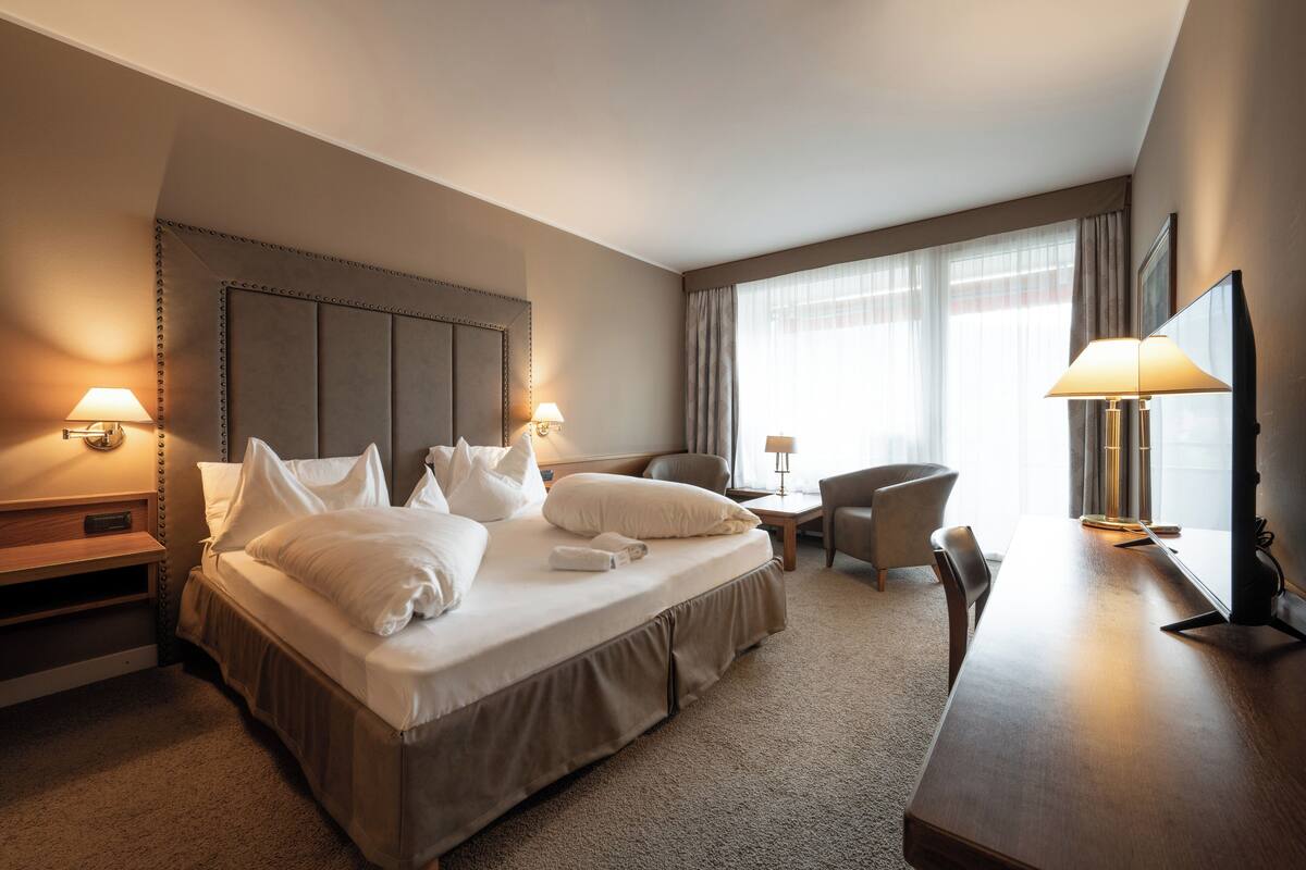 A hotel room with a large bed, two bedside tables with lamps, a desk with a chair, and a large window with curtains.