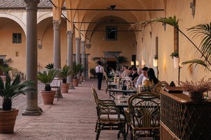 Area esterna con zona pranzo, tavoli e sedie, piante in vaso e pavimento piastrellato.
