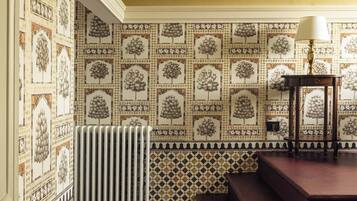 A room with patterned wallpaper, a radiator, a bench, and a table with a lamp.