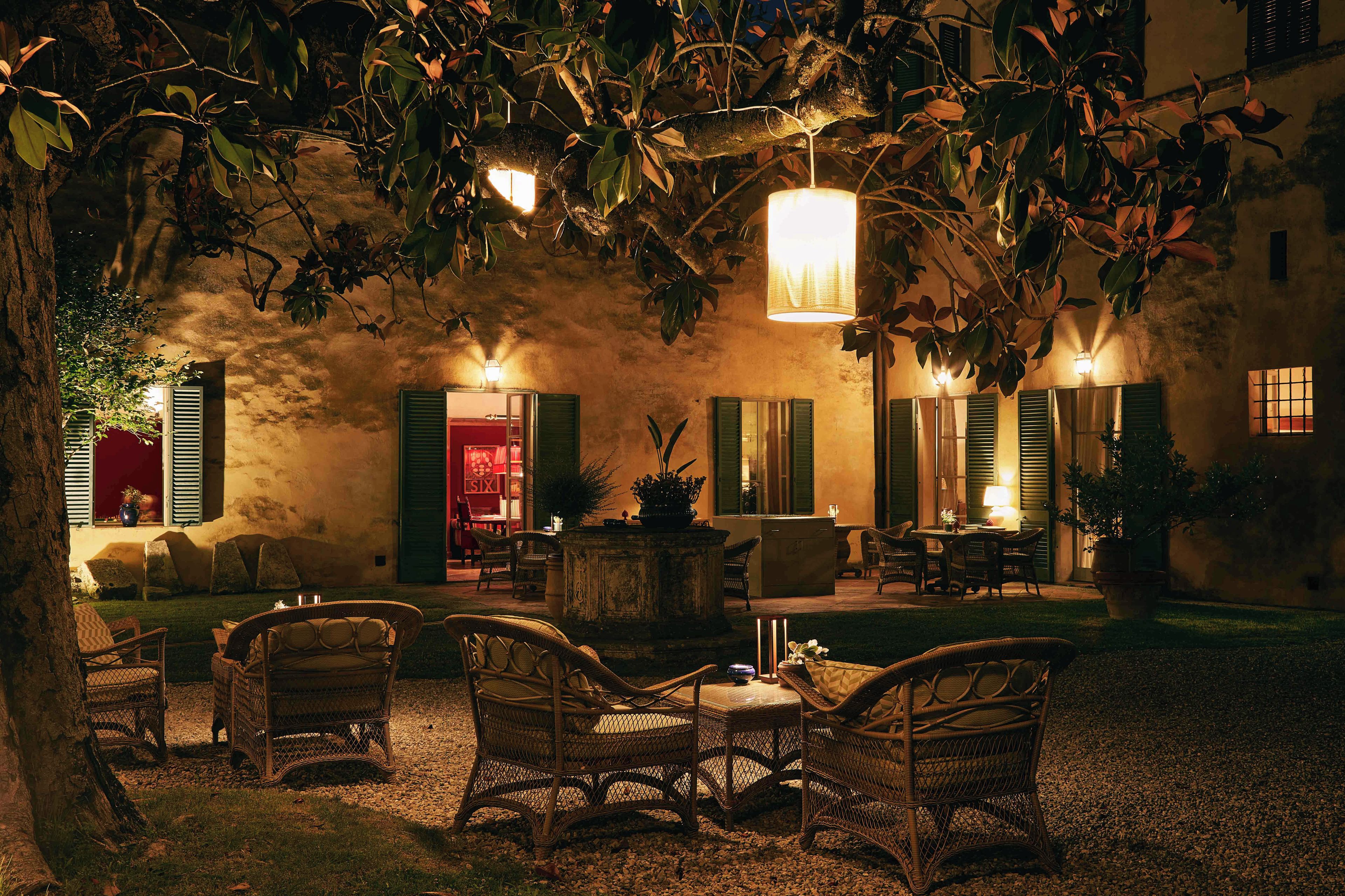An outdoor seating area with wicker furniture and a table, surrounded by greenery and a building with green shutters.