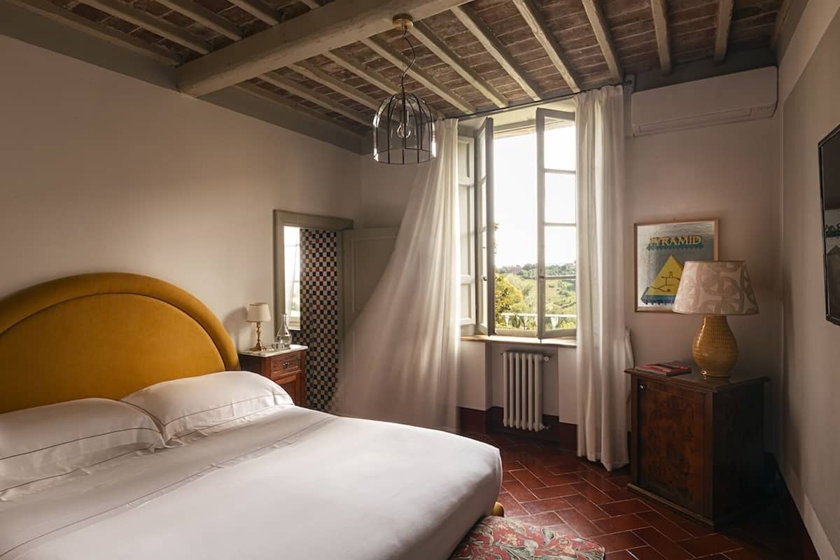 Un dormitorio con cama, ventana con cortinas, radiador, lámpara y un cuadro en la pared.