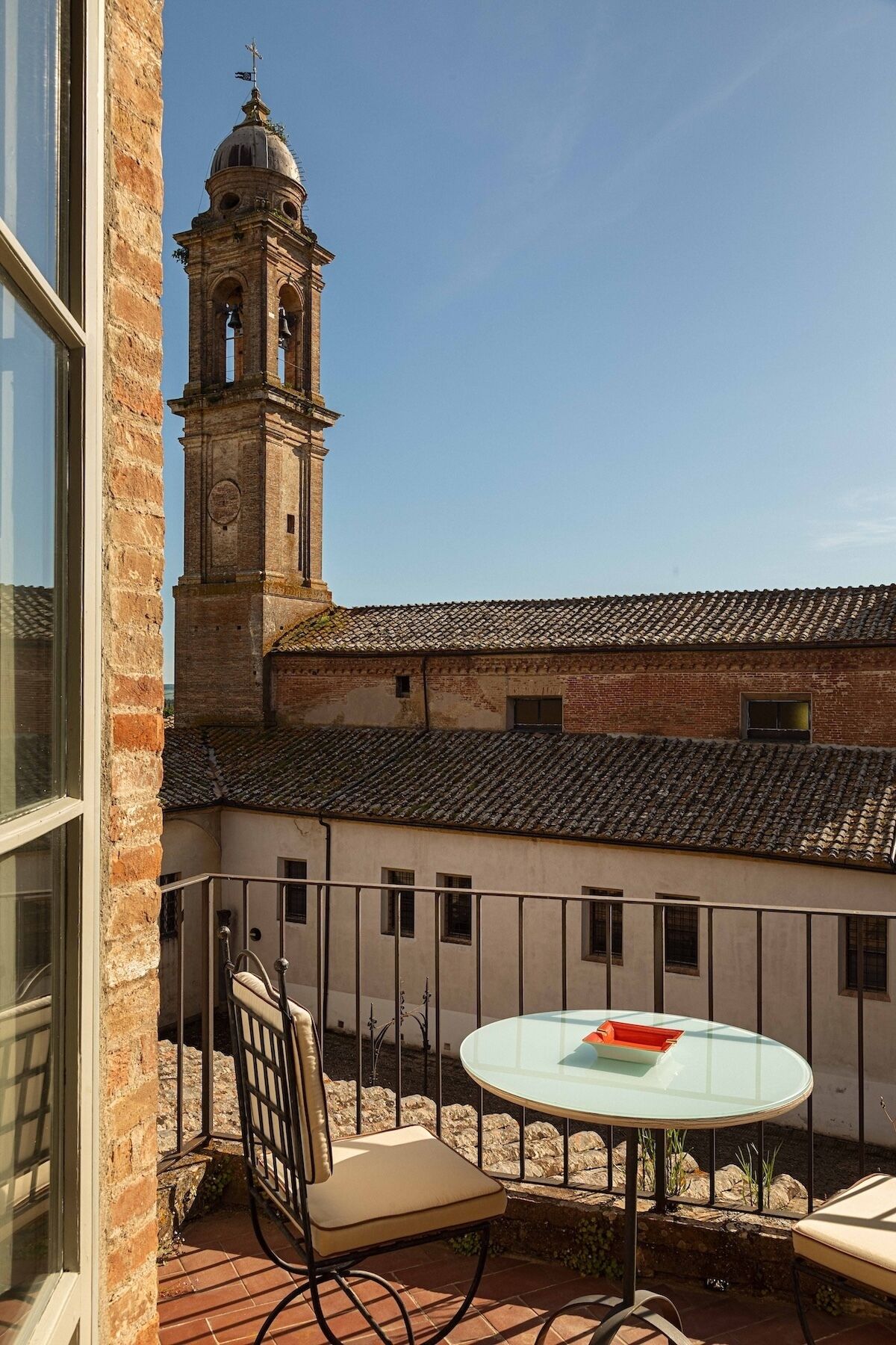 Un balcone con tavolo e sedia, affaccia su un edificio con un campanile e un tetto in tegole.