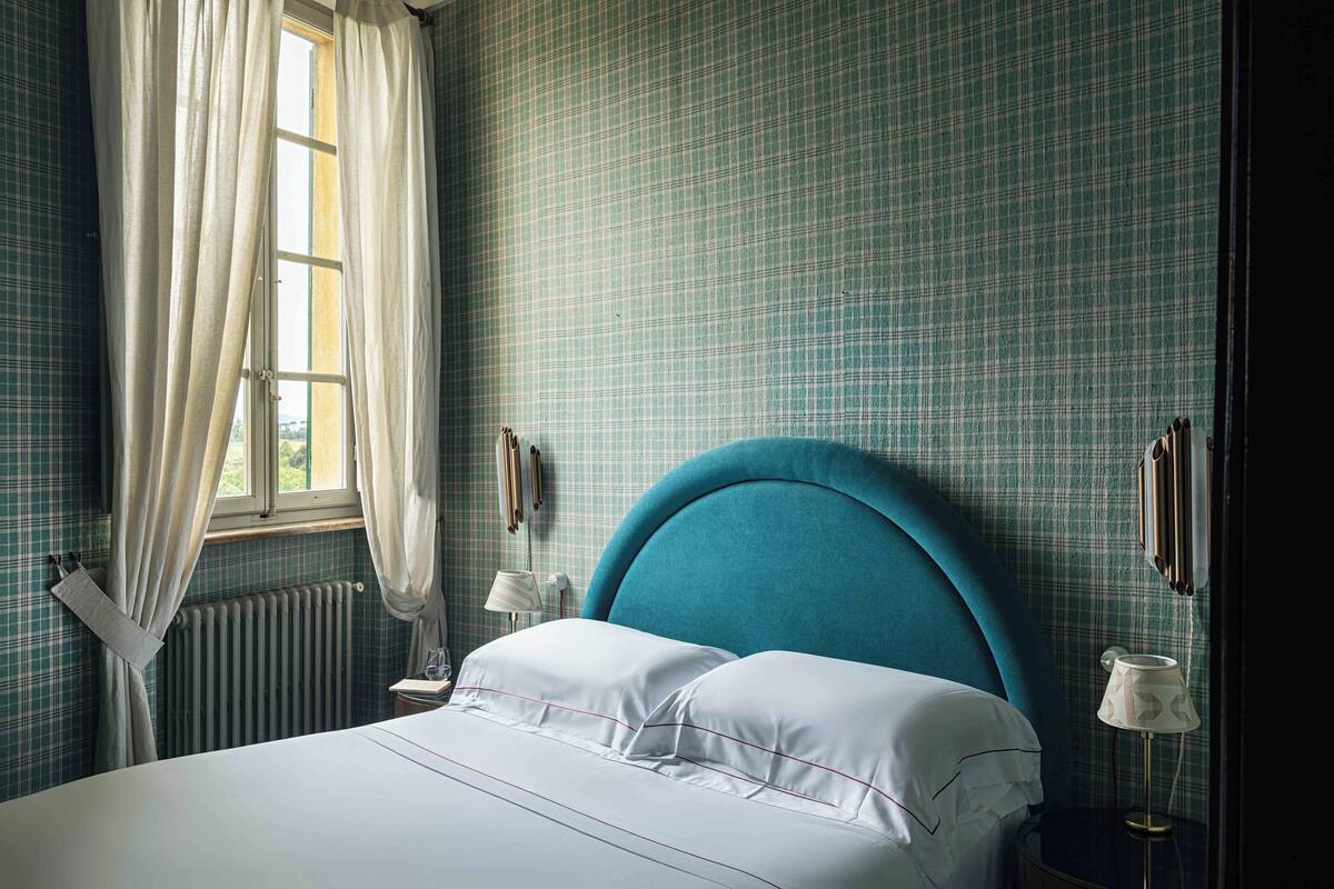 Un dormitorio con cama, cortinas, un radiador, una mesita de noche y una lámpara.