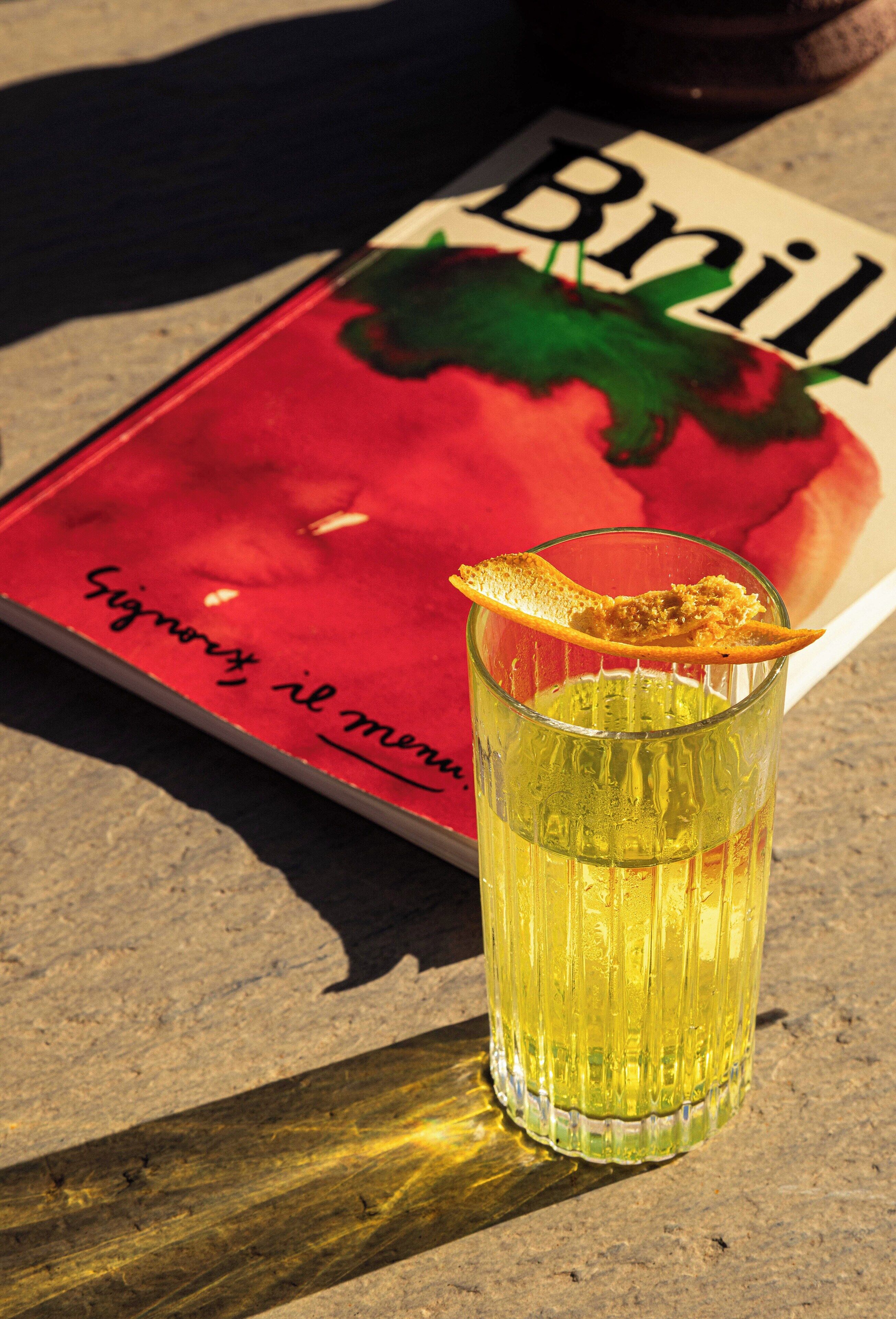 Et glas gul væske med et citronskive ved siden af en rød bog med titlen "Brill" på et bord.