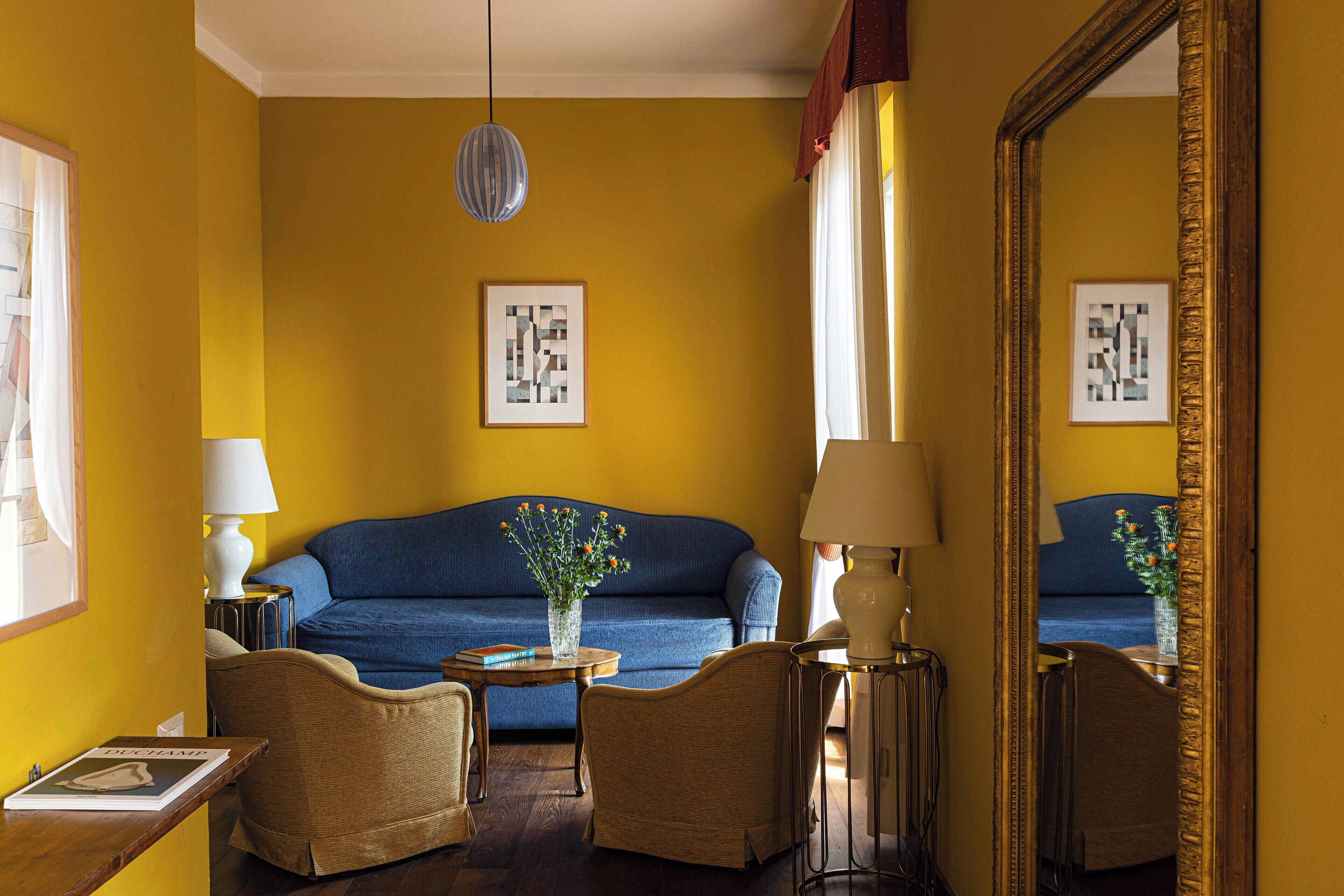 A room with a blue sofa, two wicker chairs, a small table with a vase, and a large mirror.