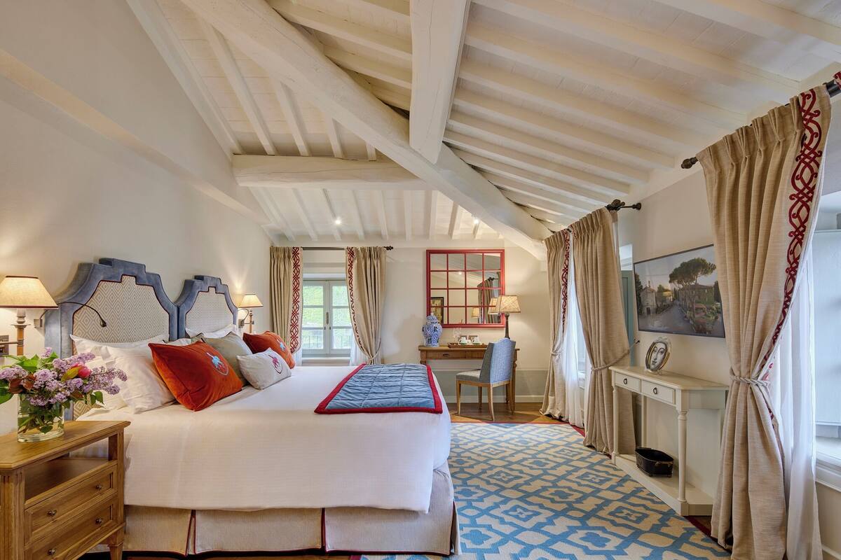 Ein Schlafzimmer mit einem großen Bett, einem Schreibtisch und einem Stuhl. Ein Fenster mit Vorhängen, ein Spiegel und ein Bild sind ebenfalls vorhanden.