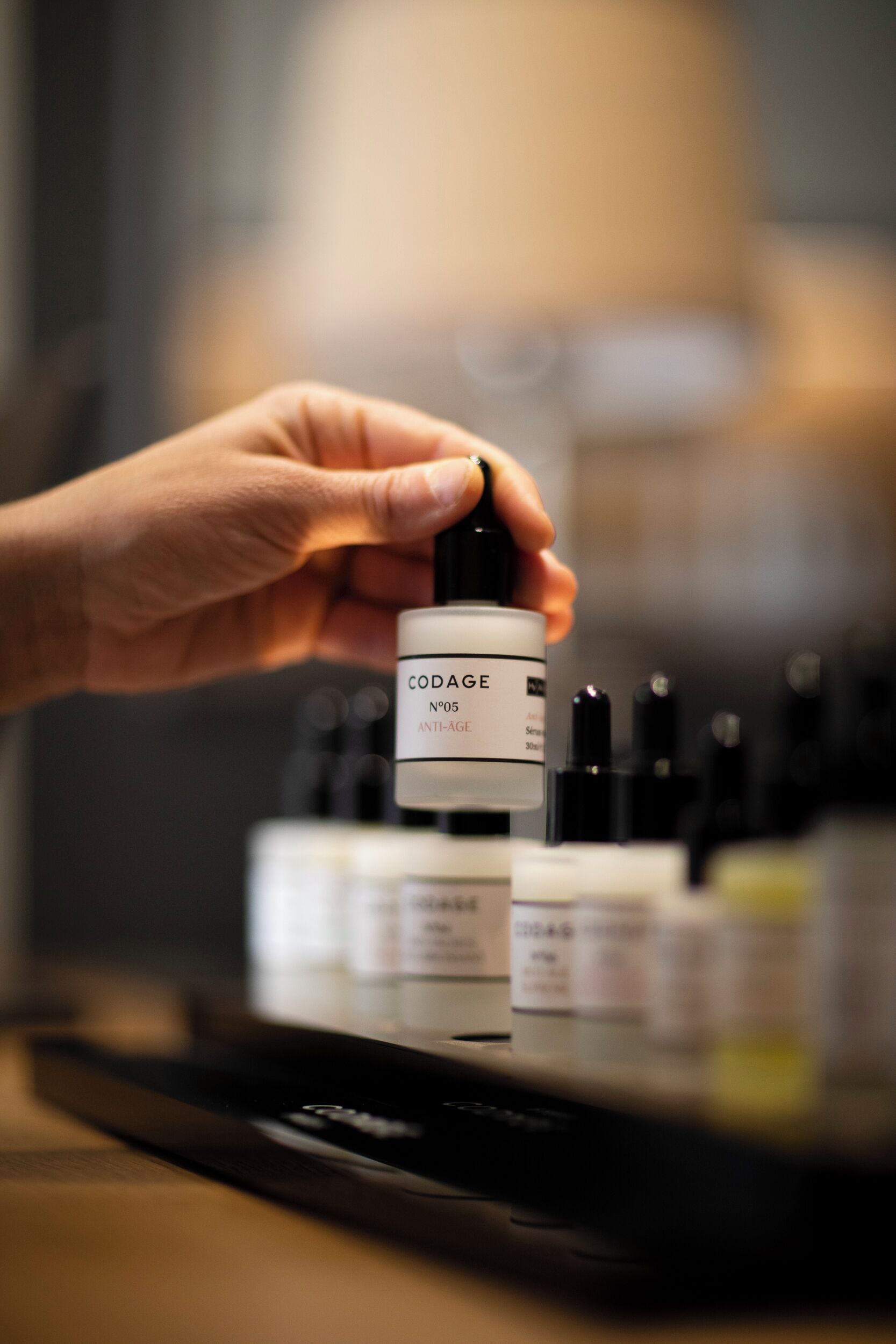 Eine Hand hält eine Flasche mit dem Aufdruck "CODAGE N°05 ANTI-AGE", im Hintergrund sind weitere ähnliche Flaschen zu sehen.