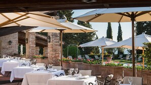 Un espace extérieur aménagé pour les repas, avec des tables pour deux personnes, protégées par de grands parasols, et un mur de briques en arrière-plan.