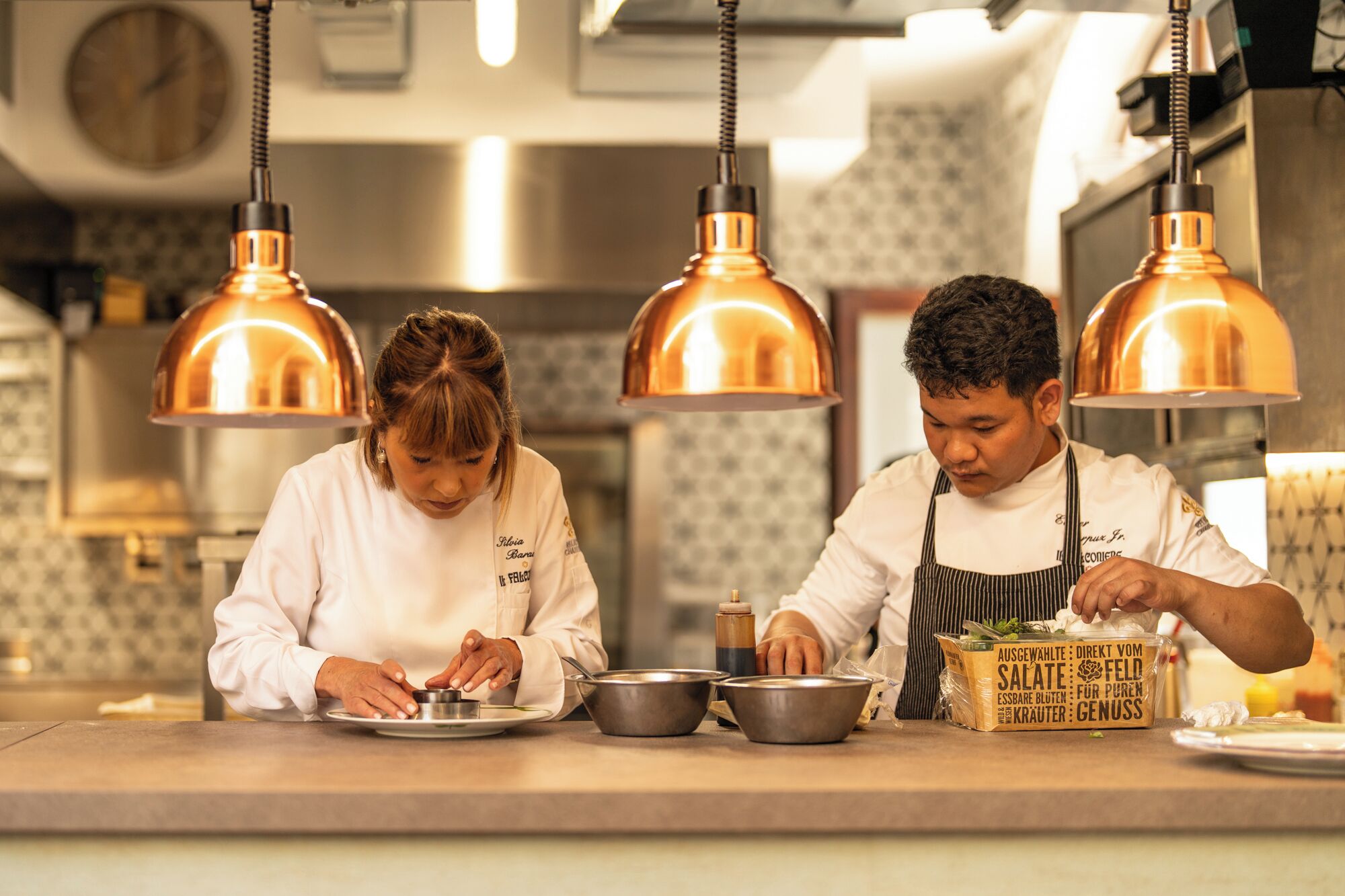 Two chefs preparing food in a kitchen with copper pendant lights and a clock on the wall.