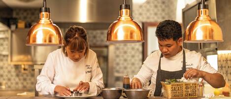 Two chefs preparing food in a kitchen with copper pendant lights and a clock on the wall.
