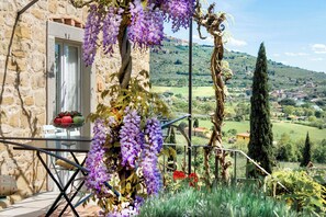 A stone building with a glass table and chairs, surrounded by purple flowers and greenery, overlooking a rural landscape with hills and trees. - Il Falconiere - Relais e Chateaux (Cortona)