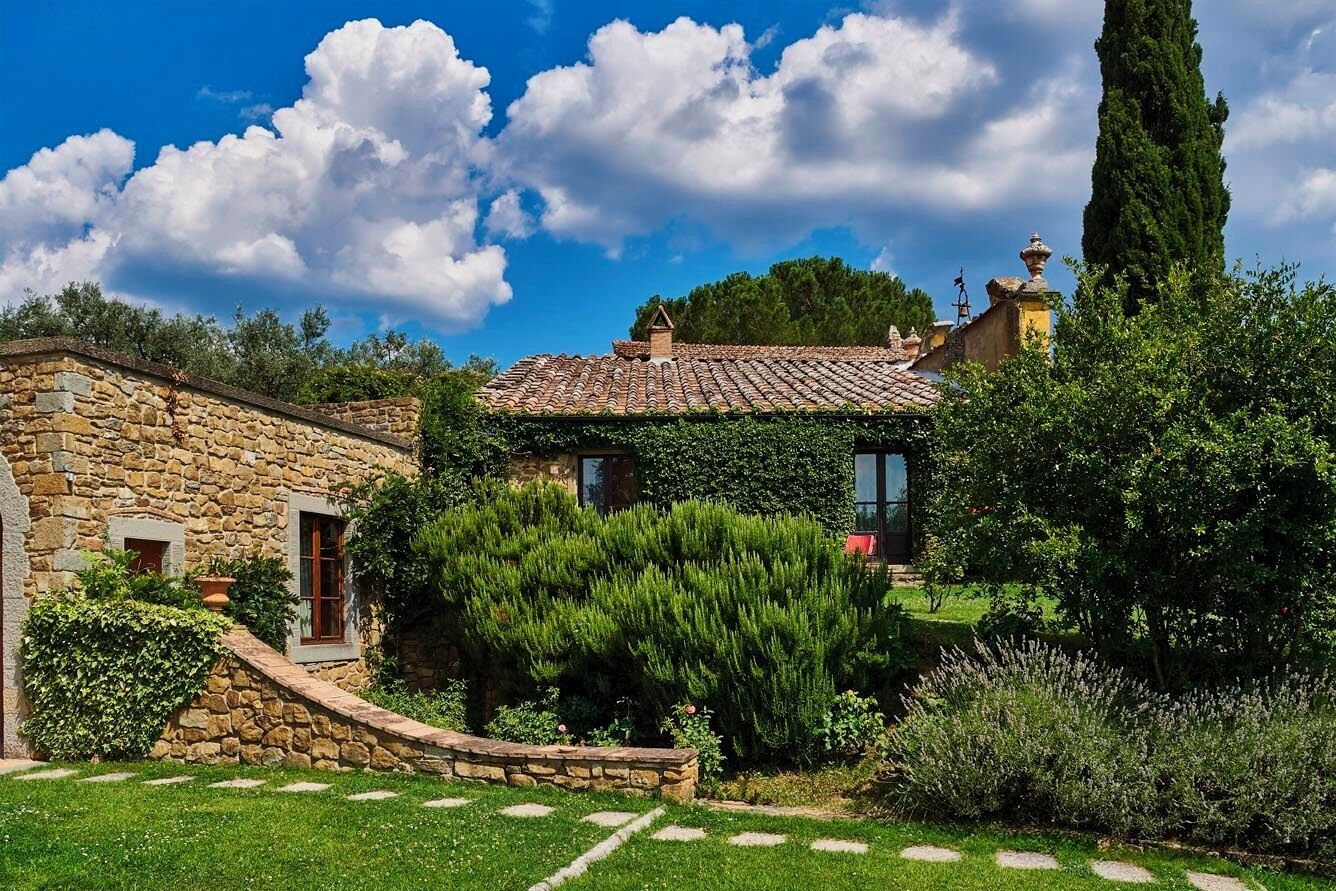A stone house with a tiled roof, surrounded by a well-maintained garden with a variety of plants and a clear blue sky.