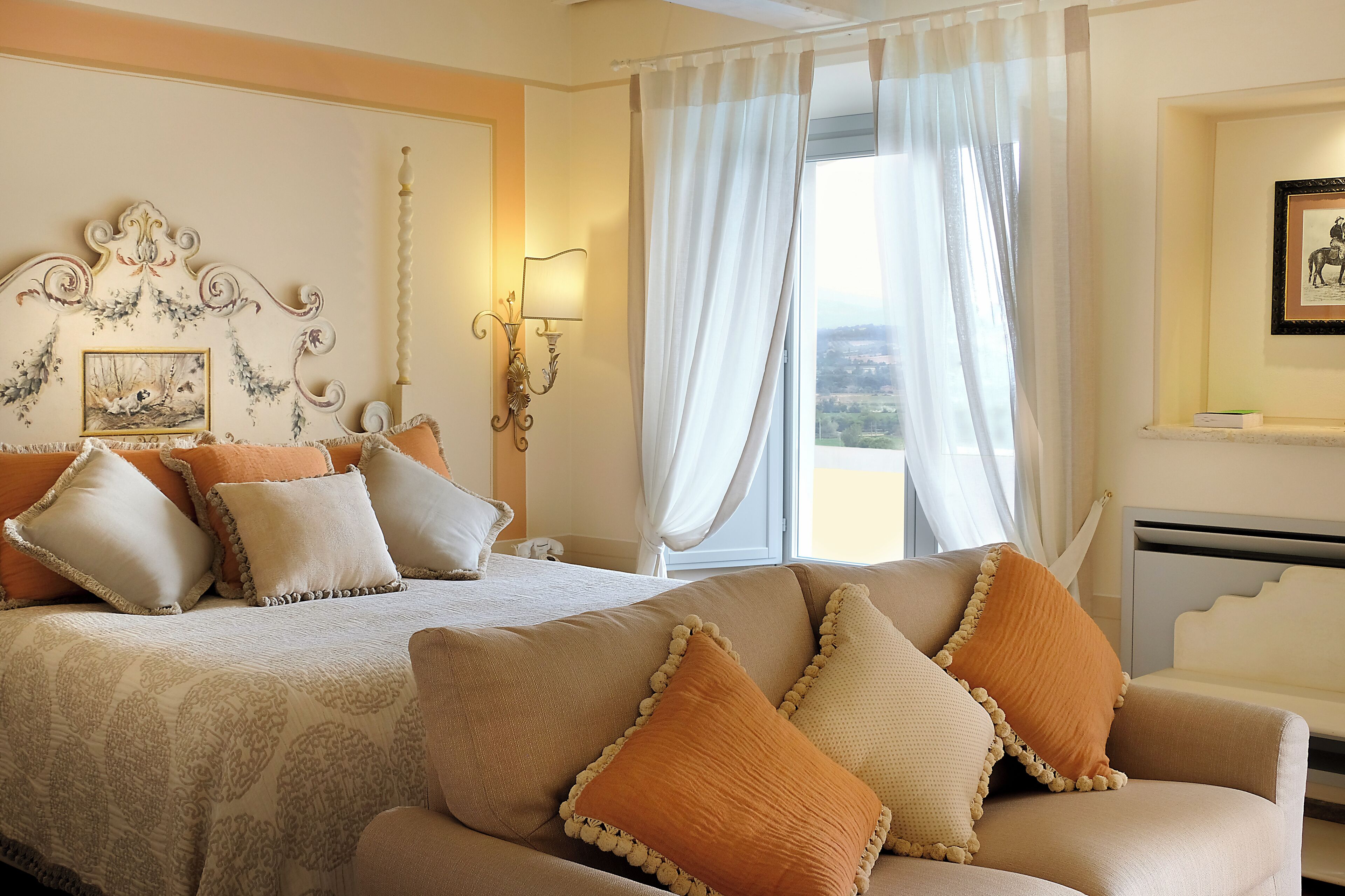 A hotel room with a bed, a sofa, a decorative headboard, a wall-mounted lamp, and a window with curtains.