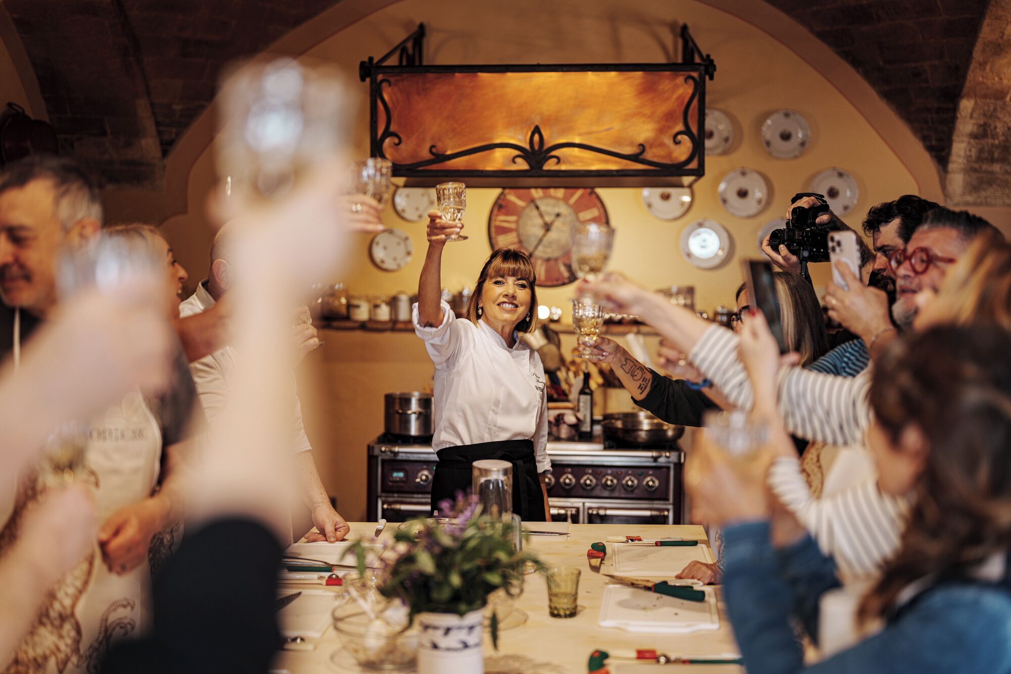 A group of people toasting with drinks in a warmly lit room with a stone ceiling and a large clock on the wall.