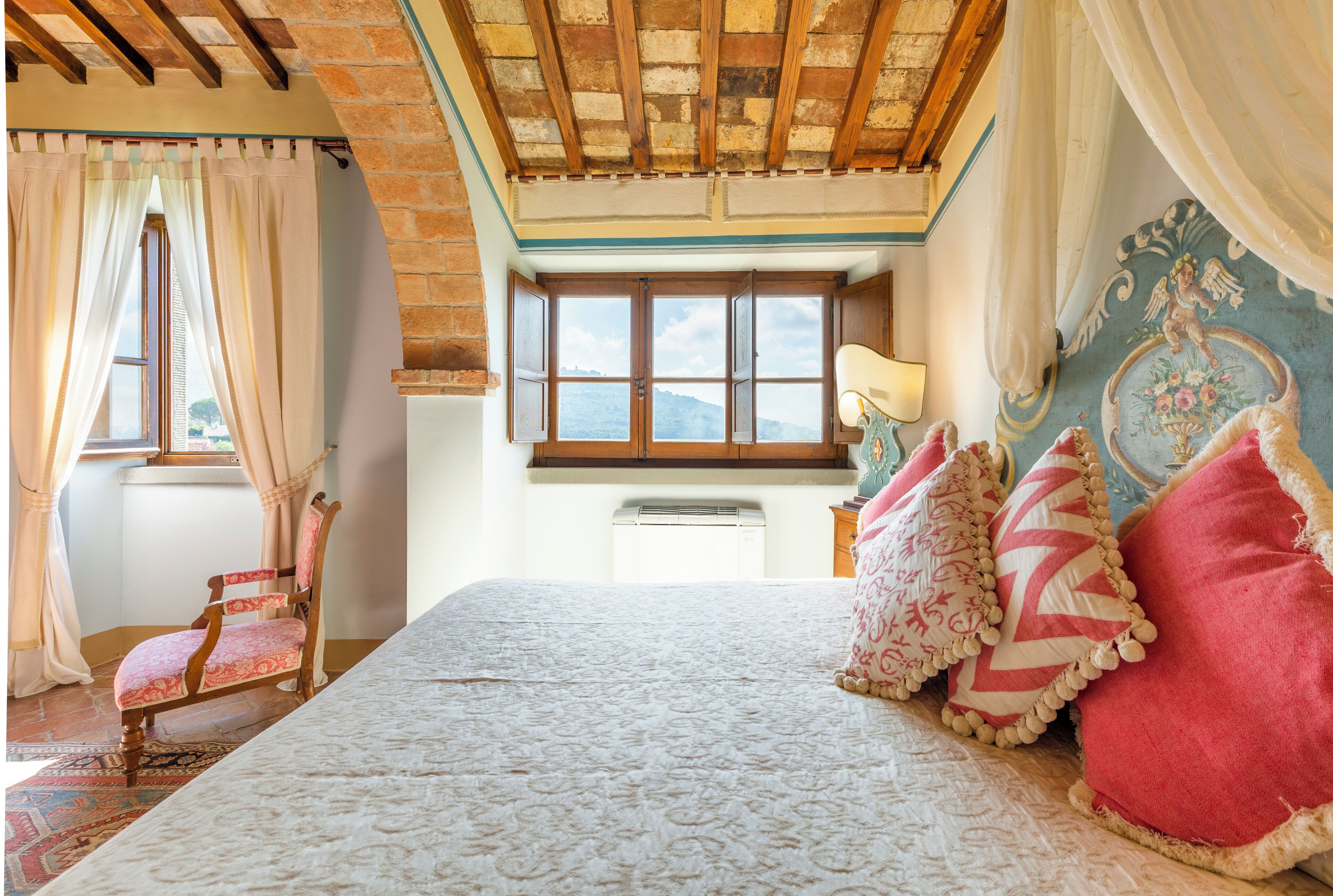 A bedroom with a large bed, a wooden chair, a window with a view, and a decorative wall.
