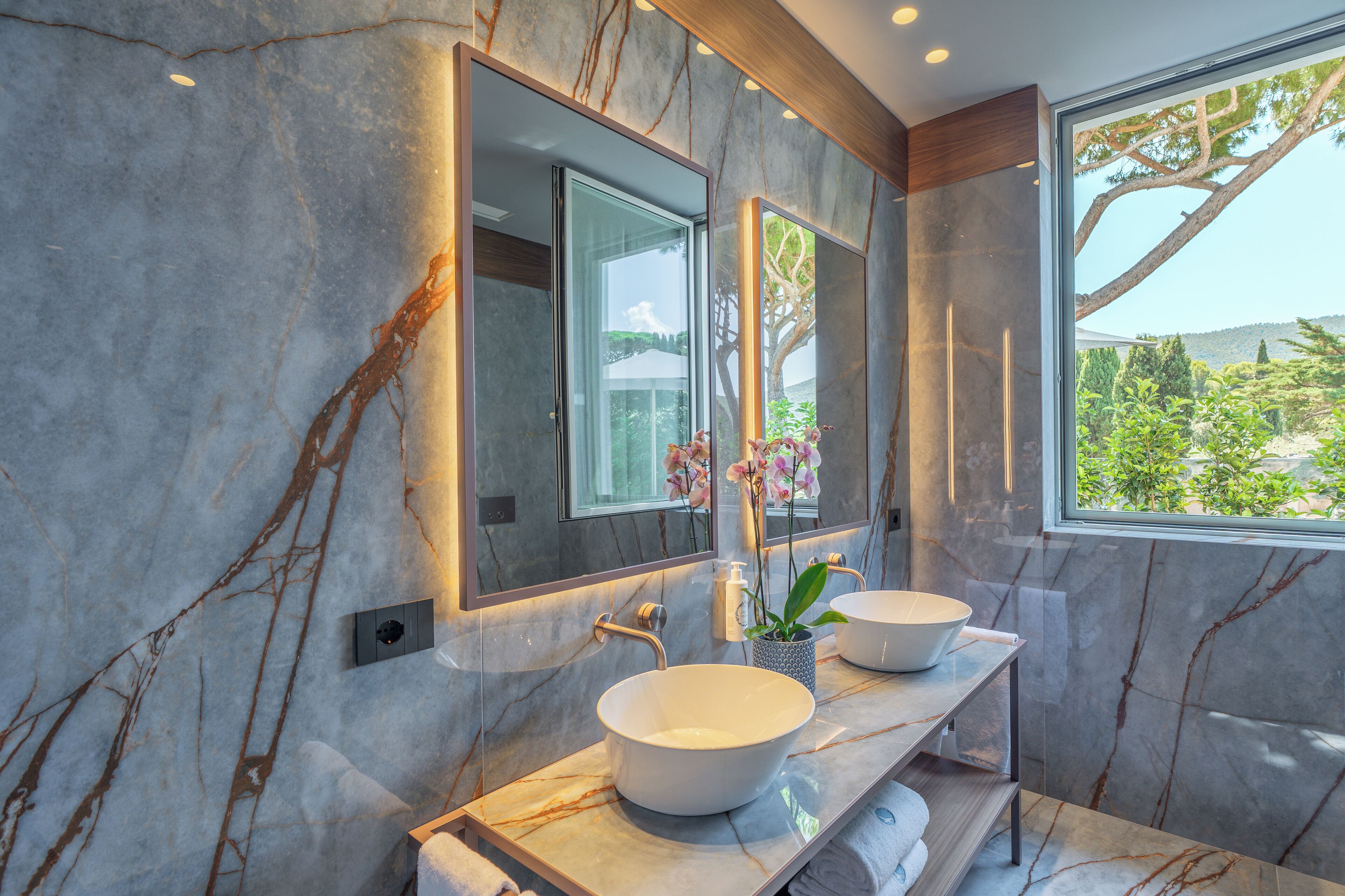 Ein modernes Badezimmer mit zwei Waschbecken, einem großen Spiegel und Blick auf Grünfläche.